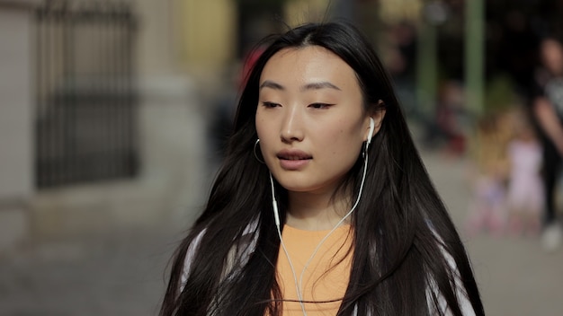 Nahaufnahme einer lächelnden asiatischen Frau, die die zentrale Stadtstraße entlang geht und Musik im Kopfhörer hört, verwenden Sie das urbane Lifestyle-Sonnenlichtkonzept des Smartphones