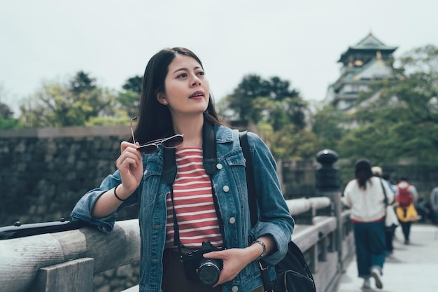 Nahaufnahme einer lächelnden jungen schönen asiatischen frau, die beiseite schaut und die kamera hält, während sie in japan reist. Fotografin stützt sich auf Geländer und steht draußen auf Brücke. Schloss Osaka im Hintergrund.