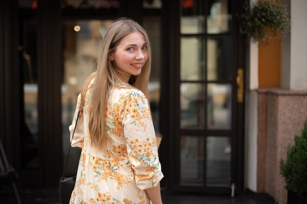 Nahaufnahme einer lächelnden kaukasischen Blondine in einem Kleid, die über ihre Schulter vor einem schwarzen Glashintergrund in der Stadt schaut