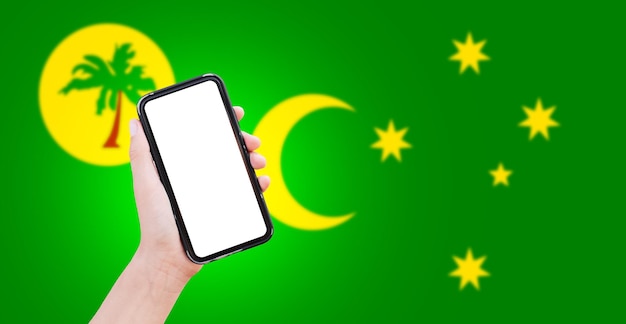 Nahaufnahme einer männlichen Hand, die ein Smartphone mit leerem Bildschirm auf dem Hintergrund der unscharfen Flagge der Cocos-Keeling-Inseln hält
