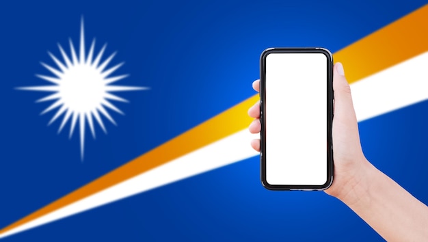 Nahaufnahme einer männlichen Hand, die ein Smartphone mit leerem Bildschirm auf dem Hintergrund der unscharfen Flagge der Marshallinseln hält