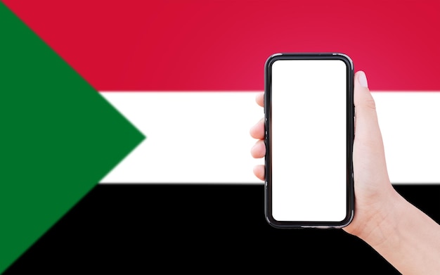 Nahaufnahme einer männlichen Hand, die ein Smartphone mit leerem Bildschirm auf dem Hintergrund der unscharfen Flagge des Sudan hält