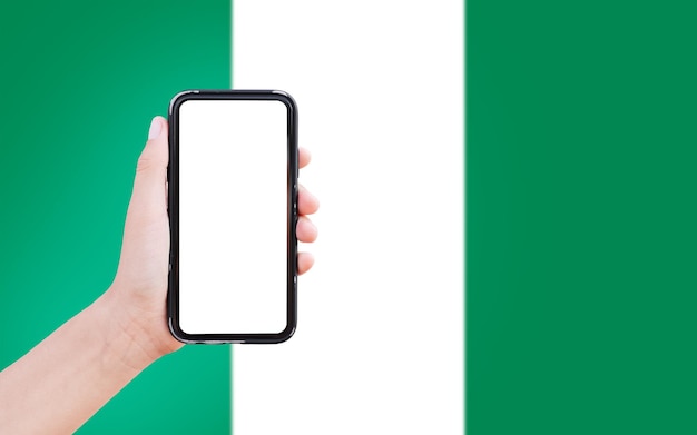 Nahaufnahme einer männlichen Hand, die ein Smartphone mit leerem Bildschirm auf dem Hintergrund der unscharfen Flagge Nigerias hält