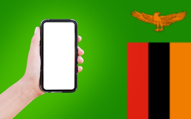 Nahaufnahme einer männlichen Hand, die ein Smartphone mit leerem Bildschirm auf dem Hintergrund der unscharfen Flagge Sambias hält