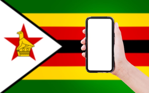 Nahaufnahme einer männlichen Hand, die ein Smartphone mit leerem Bildschirm auf dem Hintergrund der unscharfen Flagge Simbabwes hält