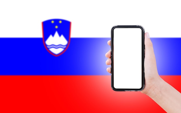 Nahaufnahme einer männlichen Hand, die ein Smartphone mit leerem Bildschirm auf dem Hintergrund der unscharfen Flagge Sloweniens hält