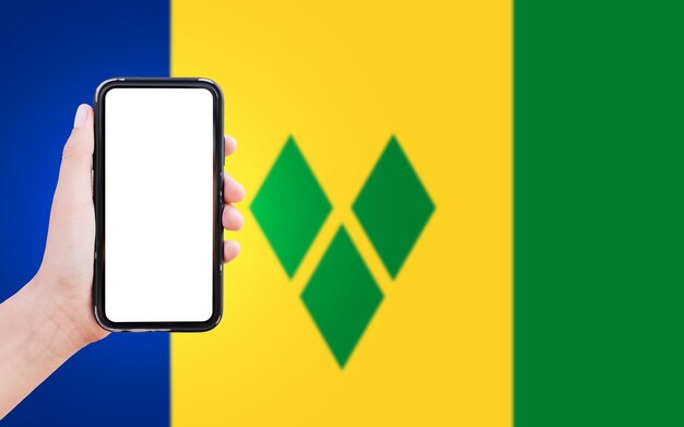 Nahaufnahme einer männlichen Hand, die ein Smartphone mit leerem Bildschirm auf dem Hintergrund der unscharfen Flagge von St. Vincent und die Grenadinen hält