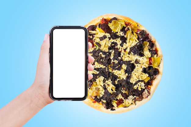 Nahaufnahme einer männlichen Hand, die ein Smartphone mit leerem Bildschirm auf dem Hintergrund einer Pizza hält, die auf blauem Hintergrund isoliert ist Veganes Lebensmittel-Lieferkonzept