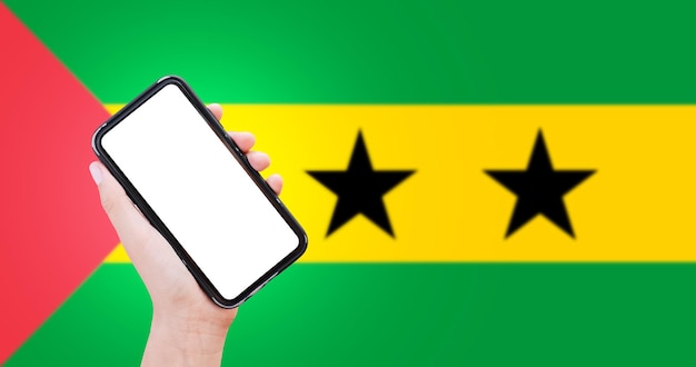 Nahaufnahme einer männlichen Hand, die ein Smartphone mit Leerzeichen auf dem Bildschirm auf dem Hintergrund der unscharfen Flagge von Sao Tome und Principe hält