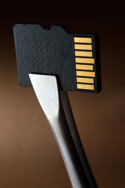 Nahaufnahme einer Micro-SD-Speicherkarte vor einem goldenen Hintergrund mit Farbverlauf, mit einer Pinzette festgeklemmt.