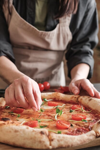 Nahaufnahme einer nicht erkennbaren Frau in Schürze, die Erbsenblätter auf Pizza mit Tomaten legt, während sie sie für die Familie zubereitet