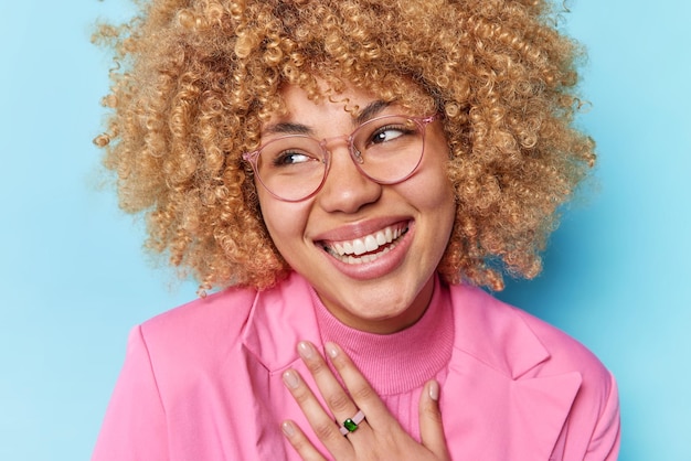 Nahaufnahme einer positiven jungen Frau mit lockigem, buschigem Haar, die fröhlich konzentriert lächelt und die Hand auf der Brust hält, eine transparente Brille und eine rosafarbene formelle Jacke isoliert auf blauem Hintergrund trägt