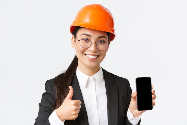 Nahaufnahme einer professionellen lächelnden Ingenieurin, Bauleiterin in Business-Anzug und Schutzhelm, mit Smartphone-Bildschirm und Daumen nach oben in Genehmigung, weißer Hintergrund
