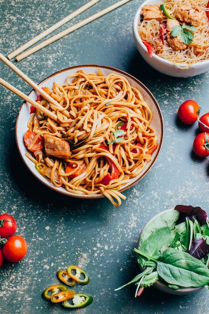 Nahaufnahme einer Schale mit chinesischen Nudeln mit Fleisch und Gemüse in Tellern und chinesischen Essstäbchen auf einer grünen Oberfläche