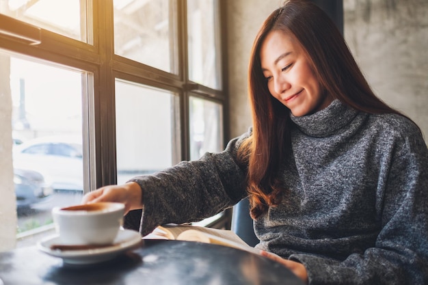 Nahaufnahme einer schönen asiatischen Frau, die sich eine Kaffeetasse schnappt, während sie ein Buch liest