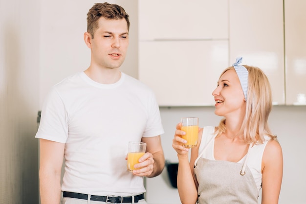 Nahaufnahme einer schönen blonden kaukasischen Frau mit einem Freund, der ein Glas mit frisch gepresstem Orangensaft hält, ein leckeres Getränk in der hellen Küche zu Hause genießt und mit Blick in die Kamera lächelt.