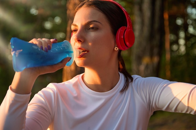 Nahaufnahme einer schönen Fitnesssportlerin in roten drahtlosen Kopfhörern, die nach dem Training Wasser aus einer wiederverwendbaren weichen Sportflasche trinkt