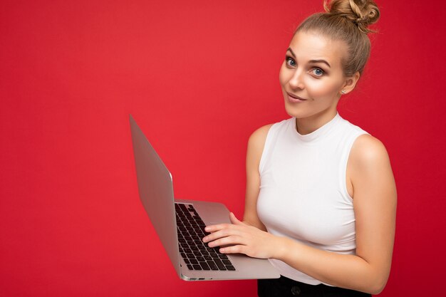 Nahaufnahme einer schönen fragenden blonden jungen Studentin mit gerafftem Haar, die ein weißes T-Shirt trägt und einen Computer-Laptop hält, der auf der Tastatur tippt und die Kamera einzeln über rotem Wandhintergrund betrachtet