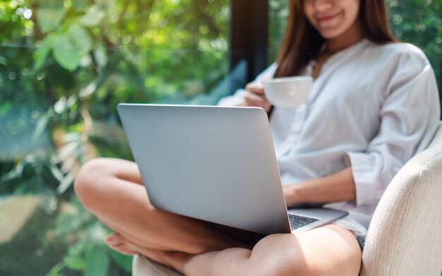 Nahaufnahme einer schönen Frau, die auf Laptop-Computer arbeitet und tippt, während Kaffee trinkt und auf einem weißen Sessel zu Hause mit grüner Natur sitzt