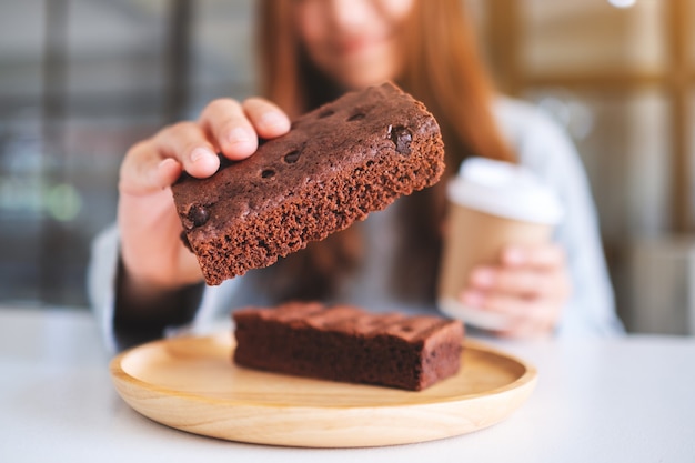 Nahaufnahme einer schönen Frau, die ein Stück Brownie-Kuchen hält und isst, während sie Kaffee trinkt