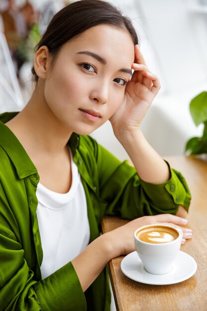 Nahaufnahme einer schönen jungen asiatischen Frau, die Kaffee trinkt, während sie im Café drinnen sitzt?