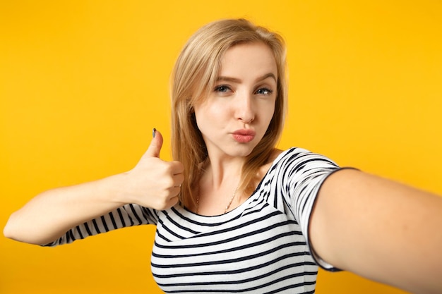 Nahaufnahme einer Selfie-Aufnahme einer schönen jungen Frau in gestreifter Kleidung, die den Daumen isoliert auf gelb-orangefarbenem Wandhintergrund im Studio zeigt. Menschen aufrichtige Emotionen, Lifestyle-Konzept. Mock-up-Kopienbereich.