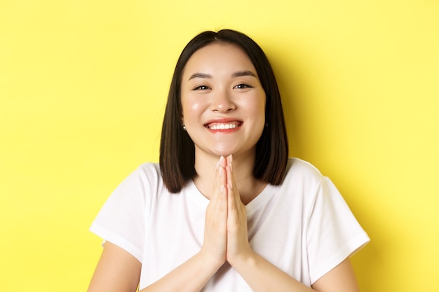 Nahaufnahme einer süßen asiatischen Frau, die Danke sagt und lächelt, Hände in Namaste hält, Geste betet und auf gelbem Hintergrund steht.