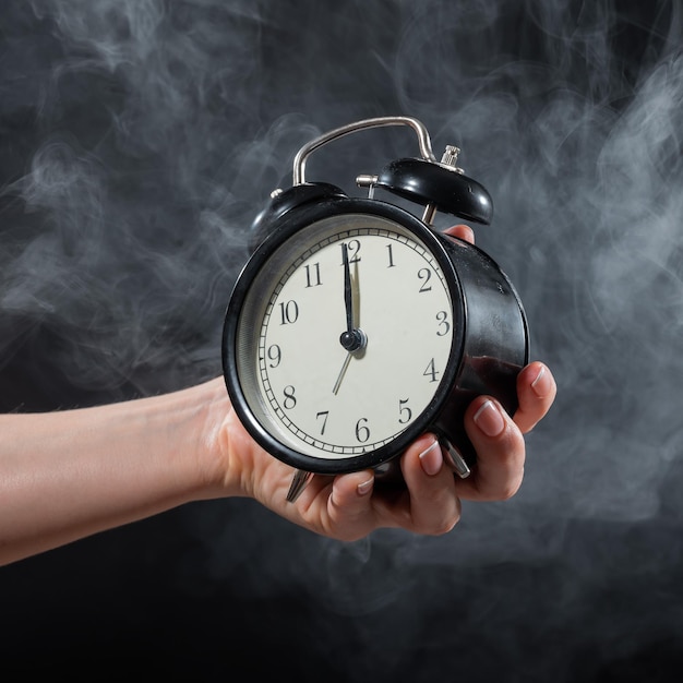 Nahaufnahme einer weiblichen Hand, die eine Uhr auf schwarzem Hintergrund in Rauch hält Wecker um Mitternacht in einem mystischen Nebel