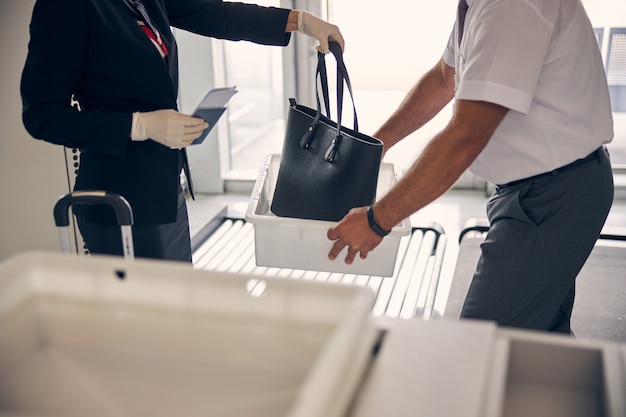 Nahaufnahme einer weiblichen Reisenden, die eine elegante Handtasche in eine weiße Box steckt, während die Flughafenmitarbeiterin einen Container für Gepäck hält