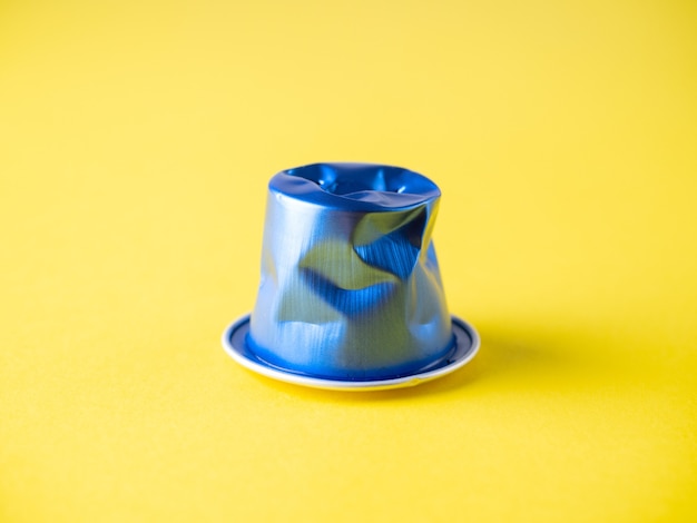 Nahaufnahme einer zerknitterten gebrauchten Aluminium-Kaffeekapsel auf gelbem Grund. Blaue Farbe, Recycling