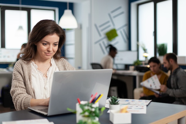 Nahaufnahme einer zufriedenen Geschäftsfrau, die die E-Mail-Eingabe auf dem Laptop beantwortet und lächelnd am Schreibtisch in einem geschäftigen Startbüro sitzt, während das vielfältige Team Statistikdaten analysiert