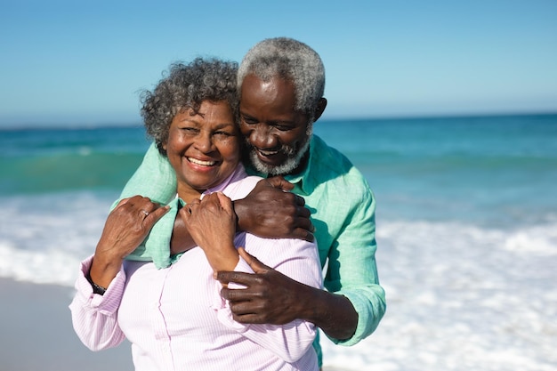 Nahaufnahme eines älteren afroamerikanischen Paares, das am Strand steht, mit blauem Himmel und Meer im Hintergrund, sich umarmt und lächelt