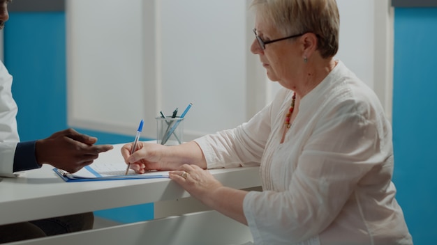 Nahaufnahme eines älteren Patienten, der Checkup-Dokumente mit Stift am Schreibtisch im medizinischen Kabinett unterschreibt. Junger Arzt, der nach einem Arzttermin eine Unterschrift auf Papierakten von einer älteren Frau benötigt