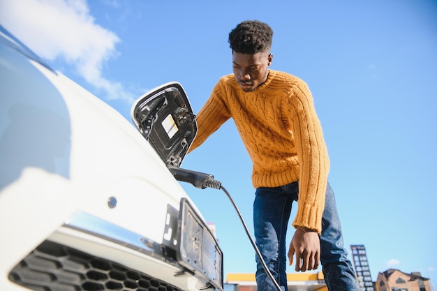 Nahaufnahme eines Afroamerikaners, der das Ladekabel an das Elektroauto anschließt. Junger Mann, der in der Nähe seines modernen Autos mit Lederkoffer in der Hand steht.