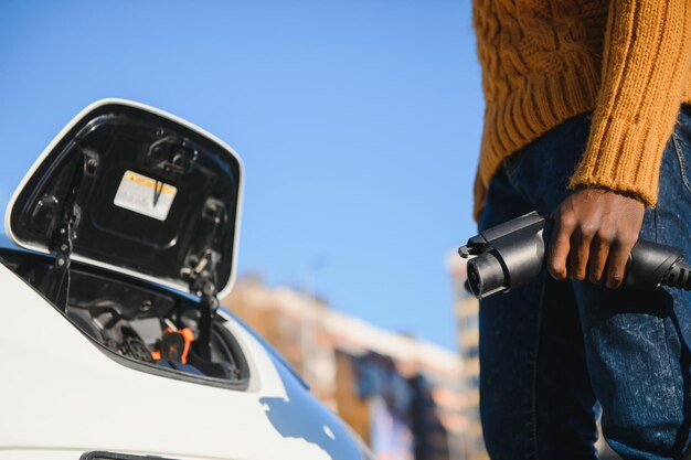 Nahaufnahme eines Afroamerikaners, der das Ladekabel an das Elektroauto anschließt. Junger Mann steht in der Nähe seines modernen Autos mit Lederkoffer in der Hand.