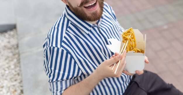 Nahaufnahme eines amüsanten jungen Hipster-Kerls, der chinesische Nudeln mit hölzernen Essstäbchen isst, die in einem sitzen