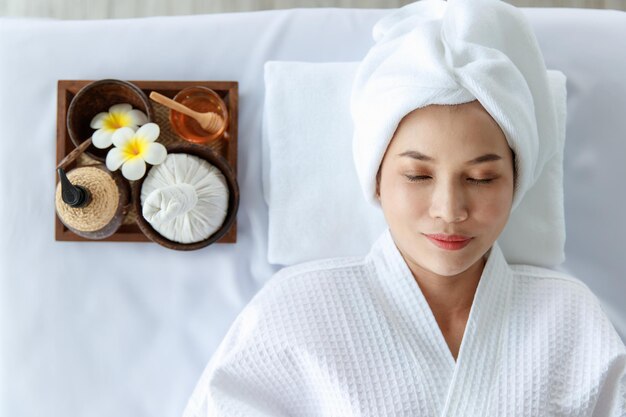 Nahaufnahme eines asiatischen, glücklichen weiblichen Modells in weißem, sauberem Bademantel, das sich mit geschlossenen Augen auf einem Massagebett mit Kompressenkugel-Kokosöl und Plumeria in einem Tablett lächelt, das auf Therapie wartet.