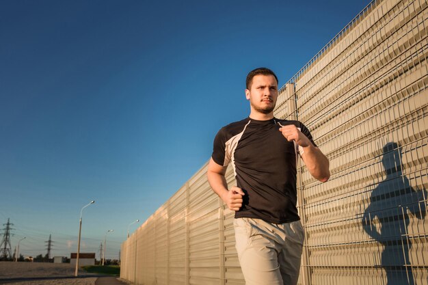 Nahaufnahme eines athletischen Mannes, der im Freien entlang einer schönen grauen Wand läuft, ein muskulöser junger Läufer, der beim Joggen im Park trainiert. Sonnenuntergang