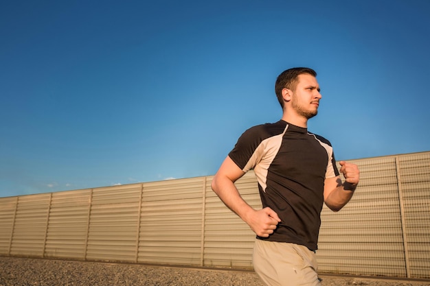 Nahaufnahme eines athletischen Mannes, der im Freien entlang einer schönen grauen Wand läuft, ein muskulöser junger Läufer, der beim Joggen im Park trainiert. Sonnenuntergang