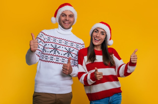 Nahaufnahme eines aufgeregten jungen verliebten Paares in Winterpullovern und Weihnachtsmützen, während sie Daumen nach oben zeigen und einzeln auf gelbem Hintergrund posieren