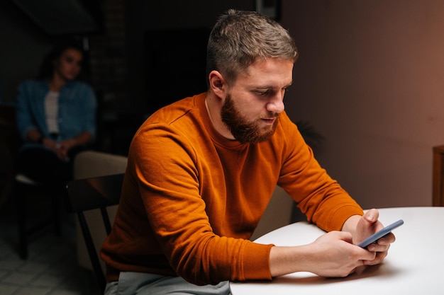 Nahaufnahme eines bärtigen jungen Mannes, der ein Handy benutzt, das am Tisch im dunklen Wohnzimmer sitzt, vor dem Hintergrund einer Freundin, die auf einem Stuhl sitzt. Hübsche männliche Schreibnachricht auf dem Smartphone zu Hause.