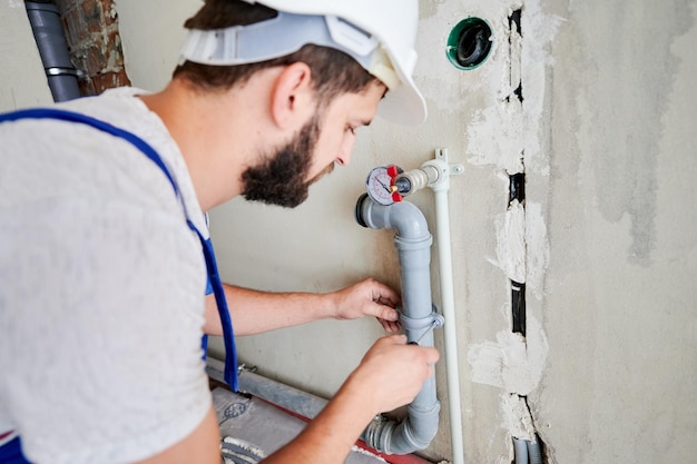 Nahaufnahme eines bärtigen jungen Mannes in Sicherheitshelm, der mit einem Schraubenzieher eine Klammer an einem Heizrohr schraubt. Mannlicher Arbeiter, der ein Wassersystem in einer Wohnung installiert. Fokus auf der Hand. Klempnerarbeiten. Hausrenovierungskonzept.