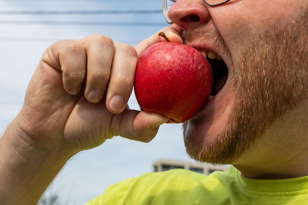 Nahaufnahme eines bärtigen Mannes im Freien, der in einen roten Apfel beißt, der gesundes Essen oder frische Produkte darstellt