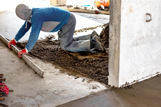 Nahaufnahme eines Baumeisters, der auf der Baustelle eine Estrichschiene auf dem mit Sandzementmischung bedeckten Boden platziert Männlicher Arbeiter, der die Oberfläche mit einer geraden Kante nivelliert, während er den Boden ebnet Verschwommener Hintergrund