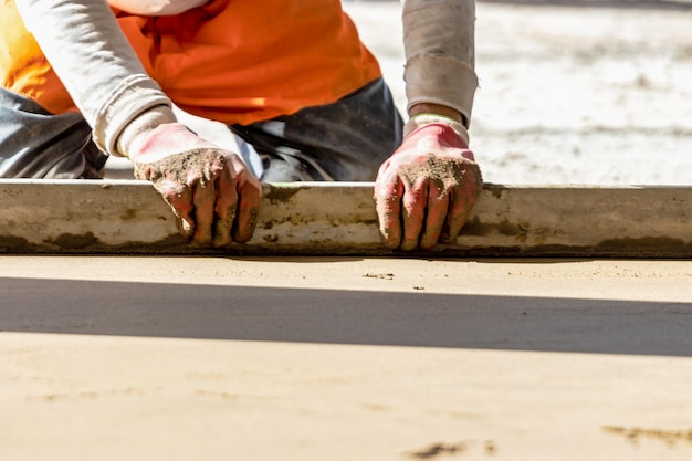 Nahaufnahme eines Baumeisters, der auf der Baustelle eine Estrichschiene auf dem mit Sandzementmischung bedeckten Boden platziert Männlicher Arbeiter, der die Oberfläche mit einer geraden Kante nivelliert, während er den Boden ebnet Verschwommener Hintergrund