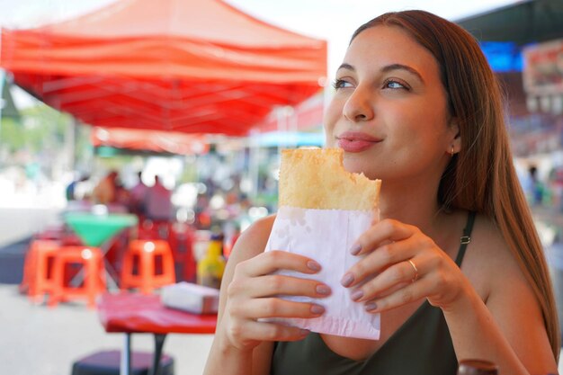 Nahaufnahme eines brasilianischen Mädchens, das auf der Messe sitzt und Pastel de Feira gefüllte gebratene Gebäck isst