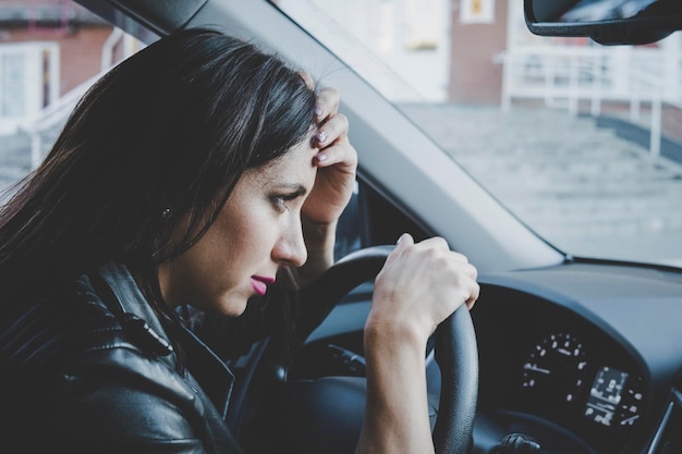 Nahaufnahme eines brünetten Mädchens, das frustriert aussieht, wenn es im Auto sitzt und das Lenkrad lehnt. Autounfall mit Fahrerin. Junge Frau fühlt sich krank oder müde, wenn sie am Steuer sitzt. Profilansicht einer besorgten Dame im Auto