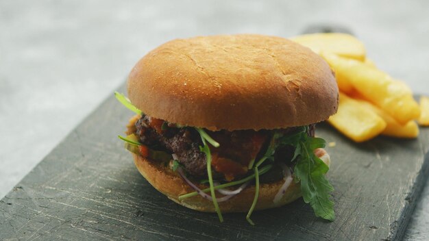 Foto nahaufnahme eines burgers mit salat und fleisch, der bei tageslicht mit goldenen pommes frites auf einem schwarzen, rauen brett serviert wird