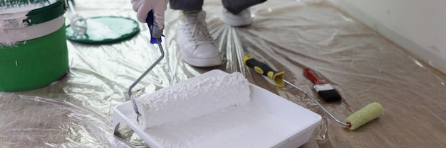 Nahaufnahme eines Dekorateurs mit Walzenpinsel, der Maler nimmt, hält Pinsel mit weißer Farbe hinein