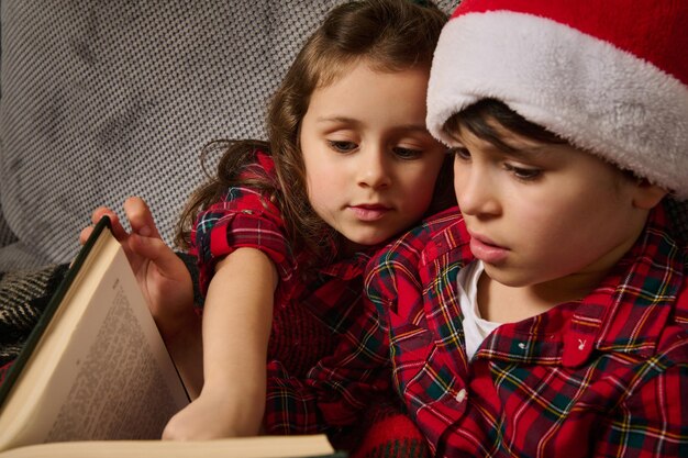 Nahaufnahme eines entzückenden kaukasischen Jungen in rot-grün kariertem Hemd und Weihnachtsmütze, der auf dem Sofa neben seiner süßen kleinen Schwester sitzt und in der Weihnachtsnacht zusammen Märchen liest. Bruder und Schwester